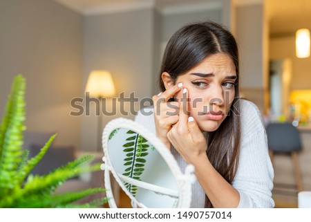 Girl looking at mirror and popping a pimple at home. Girl squeezing pimple at home. Woman examining her face in the mirror, problematic acne-prone skin concept. Upset teenager