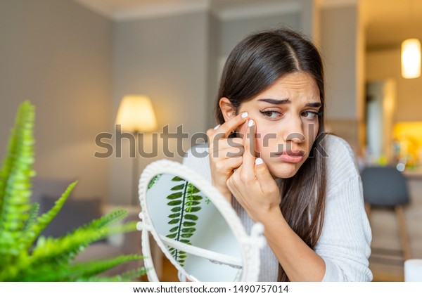 Girl looking at\
mirror and popping a pimple at home. Girl squeezing pimple at home.\
Woman examining her face in the mirror, problematic acne-prone skin\
concept. Upset teenager
