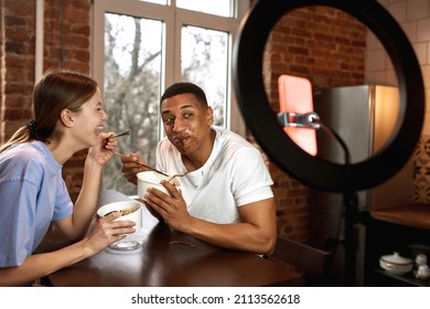 Girl Looking On Guy Face In Sauce And Laughing During Making Eat Fast Food Video Challenge On Smartphone. Multiracial Couple Of Generation Z Create Content For Social Networks At Home. Video Blogging