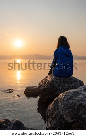 Girl looking sunset on sea, lake. Woman sitting on rocks. Lonely person. Girl alone outside. Vacation on sea. Relax sunset silhouette. Girl from behind.