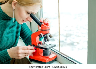 Girl Looking Through A Microscope Against The Window, To Sample Saliva, A Girl In A Green Dress With A Red Manicure. Concept Of Bacteria.