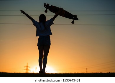 girl looks at sunset holding a longboard in her hand.