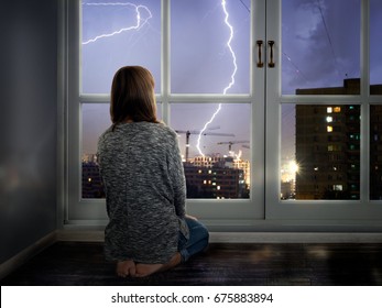 The girl looks through the window at the lightning. Thunderstorm in the city