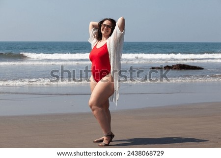 A girl with a lush sizes plus size in a red swimsuit is resting on a sandy beach against the ocean or sea