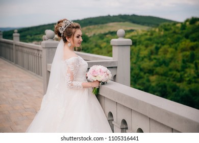 Girl in a lush wedding dress with a white bouquet of flowers is standing on a stone balcony above the precipice and admiring the view of the green mountains and fields. - Powered by Shutterstock