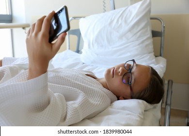 Girl lying on bed in hospital room, holding smartphone in hand and taking selfie or chatting or watching video