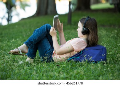 Girl Lying On Green Grass Outdoors In Summer Or Spring Reading Her Ipad Or Tablet And Wearing Headphones