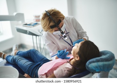 Girl Is Lying On Medical Chair In Clinic. Dentist Is Standing Above Her And Holding Mirror And Explorer While Using Binoculars