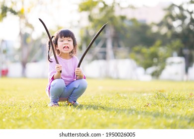 Girl Made A Cheeky Face, The Child Stuck Out Her Tongue. Child Holds The Pod Of The Golden Shower Tree. Kid Squatting In Green Lawn. During Summer Or Spring. A 4 Year Old Happy Baby Wear Pink Shirt.