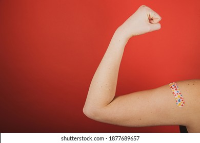 A girl makes a fist and flexes her bicep in front of a red background while wearing a bandage with hearts on it indicating she has received the Coronavirust, Covid-19 vaccination during the pandemic