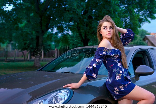 girl with make-up in blue overalls near the\
car on the background of a rural landscape, one hand on the car,\
the other holding hair, looking\
away