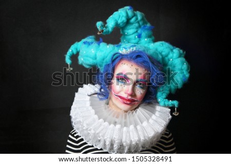 girl in makeup and costume jester . clown girl with bright makeup in blue wig On black background. She looks at the camera .