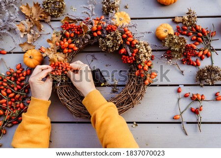 Girl making floral autumn door wreath using colorful rosehip berries, rowan, dry flowers and pumpkins. Fall flower decoration workshop, florist at work. 
