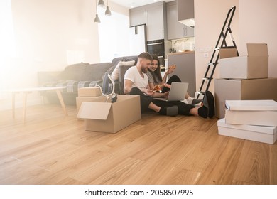 Girl and man eating pizza and using laptop at home. Young smiling european couple. Cardboard boxes with things. Concept of moving in new flat. Idea of young family. Interior of sunny studio apartment