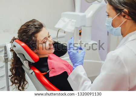 The girl in the medical chair at the dentist smiles grumbling before the treatment. A female doctor in a mask and gloves of medical instruments in his hands shows on the toes the number of teeth to be