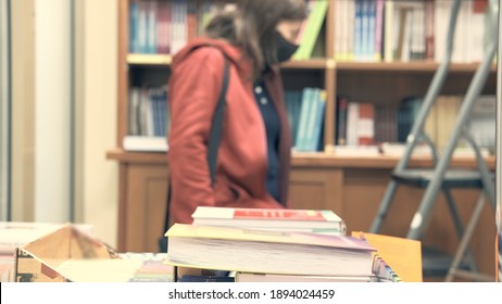 Girl in medical mask against virus, standing in library near a high bookshelf next to the stairs Camera focus on book in foreground, person is in blur