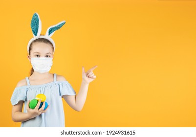 A girl in a medical mask for coronavirus on her face, on her head with bunny ears, holds Easter eggs in her hands, points to an empty space for text with her finger. Easter and spring symbol. - Shutterstock ID 1924931615