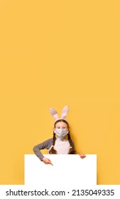 A girl with a medical mask and Easter bunny ears is holding empty white board over yellow background. Easter, coronavirus, quarantine, safe celebration.?opy space