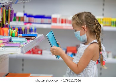 A Girl In A Medical Mask In A Shopping Center Buys Stationery For School. Preparing For School After Quarantine Covid - 19. Protective Mask For The Prevention Of Coronavirus. 