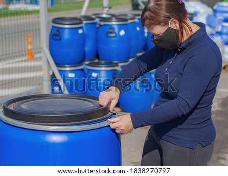 A girl in a medical mask from the virus opens a large, plastic barrel. In the background you can see pallets with plastic containers, an iron fence and a roadway for cars.