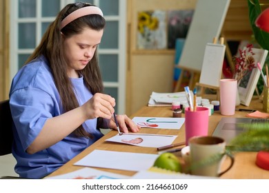 Girl With Mental Disability Painting With Watercolors And Paintbrush On Paper While Sitting By Desk In Front Of Laptop In Living Room