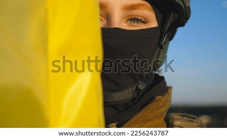 Girl in military uniform and helmet looks out from behind flag of Ukraine. Sight of female ukrainian army soldier with flag. Victory against Russian aggression. Invasion resistance concept. Close up.