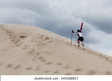 A Girl With Mountain Skis On Her Shoulders And In Ski Boots Climbs A Sand Dune. Alpine Skiing. Not The Season