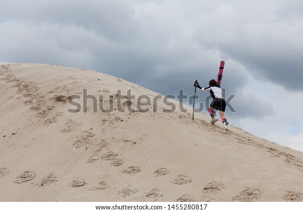 A girl with\
mountain skis on her shoulders and in ski boots climbs a sand dune.\
Alpine skiing. Not the season