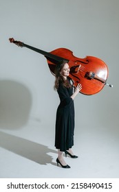 Girl musician with long hair in black dress carries the double bass against the white background.
