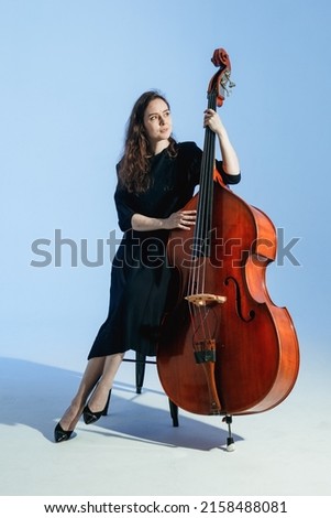 Girl musician with long hair in black dress poses with the double bass against the light blue background.