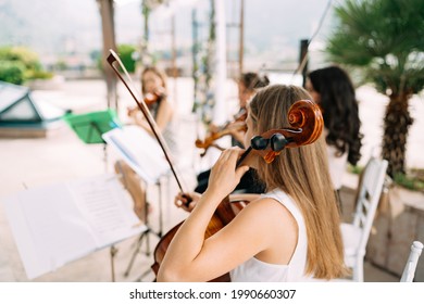 Girl Musician From The String Orchestra Sits Near The Music Stand And Holds A Cello During The Wedding Ceremony, Back View 