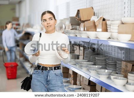 Girl near display case with kitchen utensils chooses beautiful clay plates with pattern. Customer buys view windowshopping and examines ceramic saucers and dishes