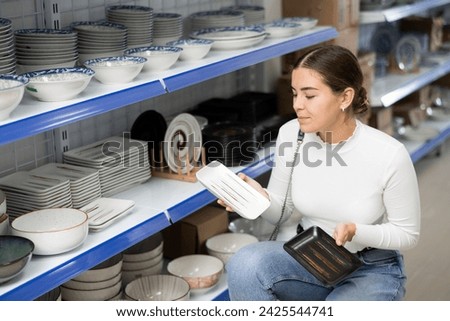 Girl near display case with kitchen utensils chooses beautiful clay square plates with pattern. Customer buys view windowshopping and examines ceramic saucers and dishes