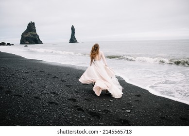 Girl In Old Old-styled Wedding Dress Are Running On The Black Sand Beach In Vic, Iceland With Rock Islands On Background.