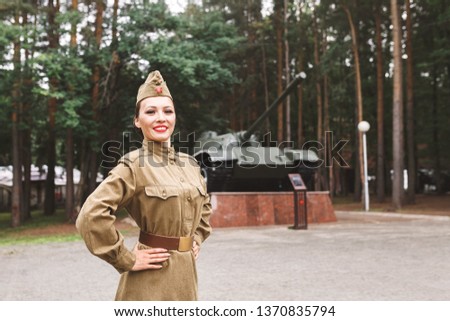 A girl in an old Soviet military uniform salute in park memorial with Tank T-34 at the background. World War II victory celebrating at 9 may in Russia