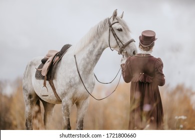 A girl in an old suit stands next to a gray horse in a harness in the middle of tall grass - back view