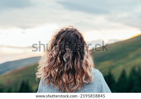 girl on the background of mountains forest and sunset looks into the distance travel hike conquering the peaks from the back fair-haired curly pleasure from life prayer look into the sky to God