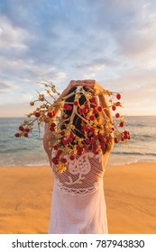 Girl On Beach In White Lace Macrame Dress Facing The Ocean At Sunset Holding Red Date Palm Frond As Head Dress