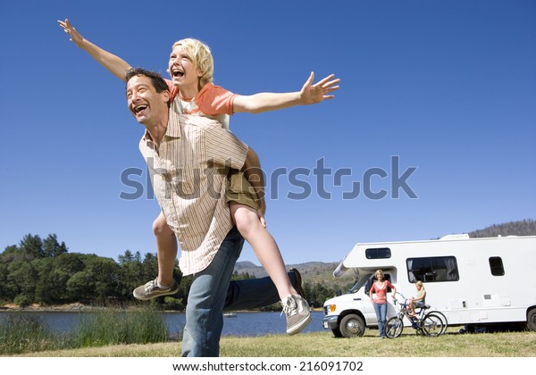 Girl on father\'s back, mother\
and brother (10-12) by motor home in background, low angle\
view
