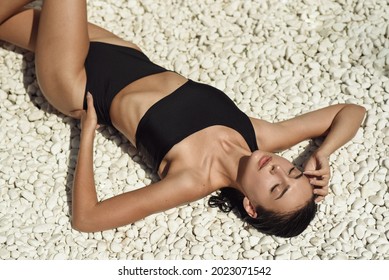 girl on a sunny day sunbathes in a designer swimsuit on the beach with pebbles and palm trees. summer rest. beautiful woman in black swimsuit in sexy pose sunbathes on the beach with white pebbles