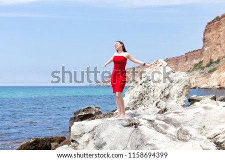 Girl on wild rocky seashore. Attractive female person in red sundress barefoot standing on rock with arms outstretched