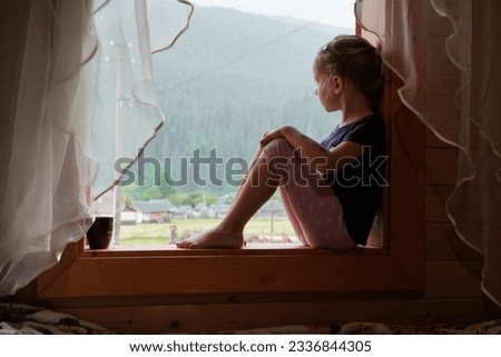 Girl on the windowsill looking on rainy mountains. Child sitting in the house and looking outdoors, waiting for nice weather, observing nature, thinking. Relaxed sad kid alone at home