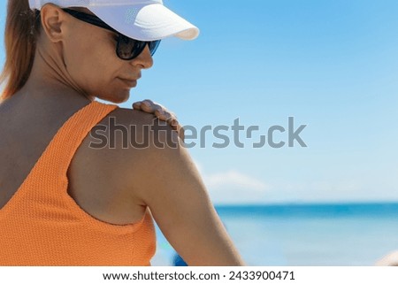 A girl in an orange bikini, white cap, and sunglasses applies sunscreen to her shoulders on the beach, epitomizing the essence of summer relaxation and sun care amidst the coastal breeze and sands. Stock photo © 