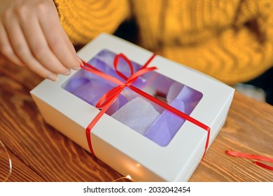 Girl In Orange Knitted Sweater Opens Festive Gift. Christmas Or Birthday. Close-up Of Hand Pulling Ribbon From Box On Wooden Table.