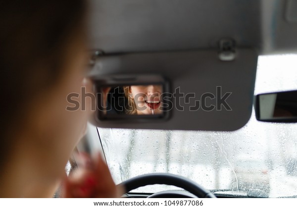The girl paints her lips with red lipstick look in\
the mirror in the sun shade in her car. A young girl puts make-up\
in the car.