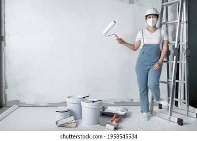 Girl Paints A White Wall With A Roller. Repair Of The Interior. Young Female Decorator Painting A Wall In The Empty Room, Concept Builder Or Painter In Helmet With Paint Roller Over The Empty Room.