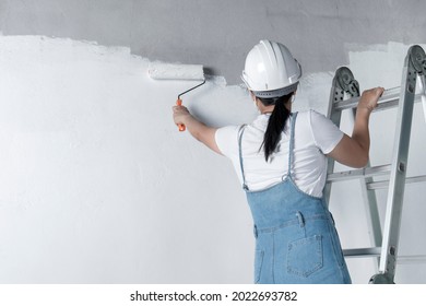 Girl Paints A White Wall With A Roller. Repair Of The Interior. Young Female Decorator Painting A Wall In The Empty Room, Concept Builder Or Painter In Helmet With Paint Roller Over The Empty Room.