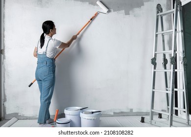 Girl Paints A White Wall With A Roller. Repair Of The Interior. Young Female Decorator Painting A Wall In The Empty Room, Concept Builder Or Painter In Helmet With Paint Roller Over The Empty Room.