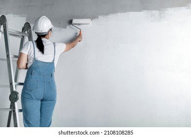 Girl Paints A White Wall With A Roller. Repair Of The Interior. Young Female Decorator Painting A Wall In The Empty Room, Concept Builder Or Painter In Helmet With Paint Roller Over The Empty Room.