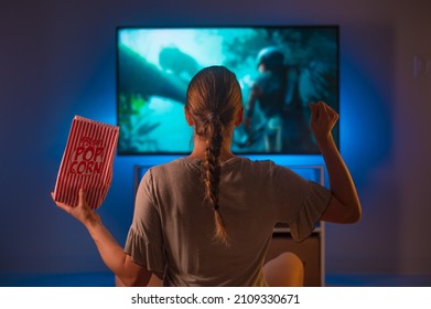 A girl in pajamas sits on the bed in the bedroom and watches TV. She has a packet of popcorn in her hand. Shooting from the back. neon light. Insomnia, watching your favorite movies and TV shows. - Shutterstock ID 2109330671